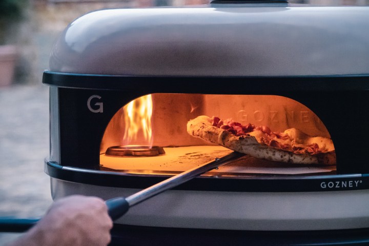 Hand placing pizza into a Gozney outdoor oven with visible flames inside.