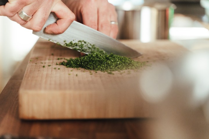 Person chopping green herbs on a wooden cutting board with a knife.