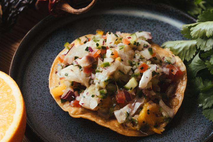 Tostada topped with diced seafood and vegetables on a dark plate, surrounded by herbs and dried peppers.