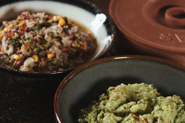 Two bowls with guacamole and salsa on a dark countertop.
