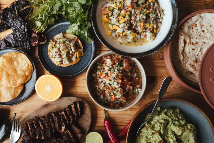 Mexican food spread with tortillas, guacamole, ceviche, salsa, grilled meat, and fresh herbs on a wooden table.