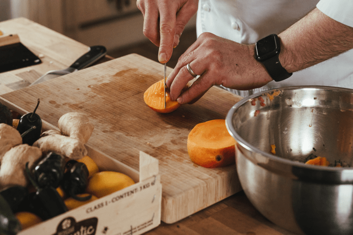 Person slicing a persimmon on a wooden board near ginger and peppers.