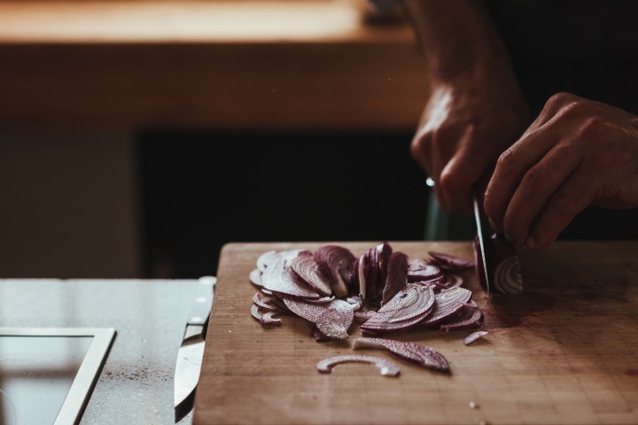 Person slicing a red onion on a wooden cutting board in a kitchen setting.