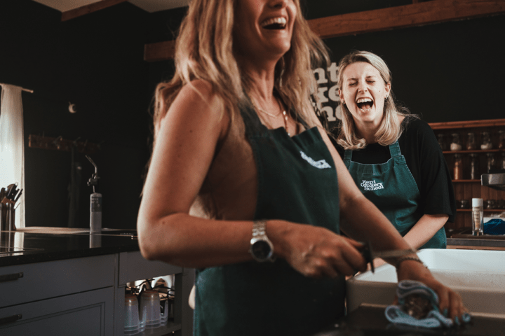 Two women in aprons laughing together in a kitchen setting.