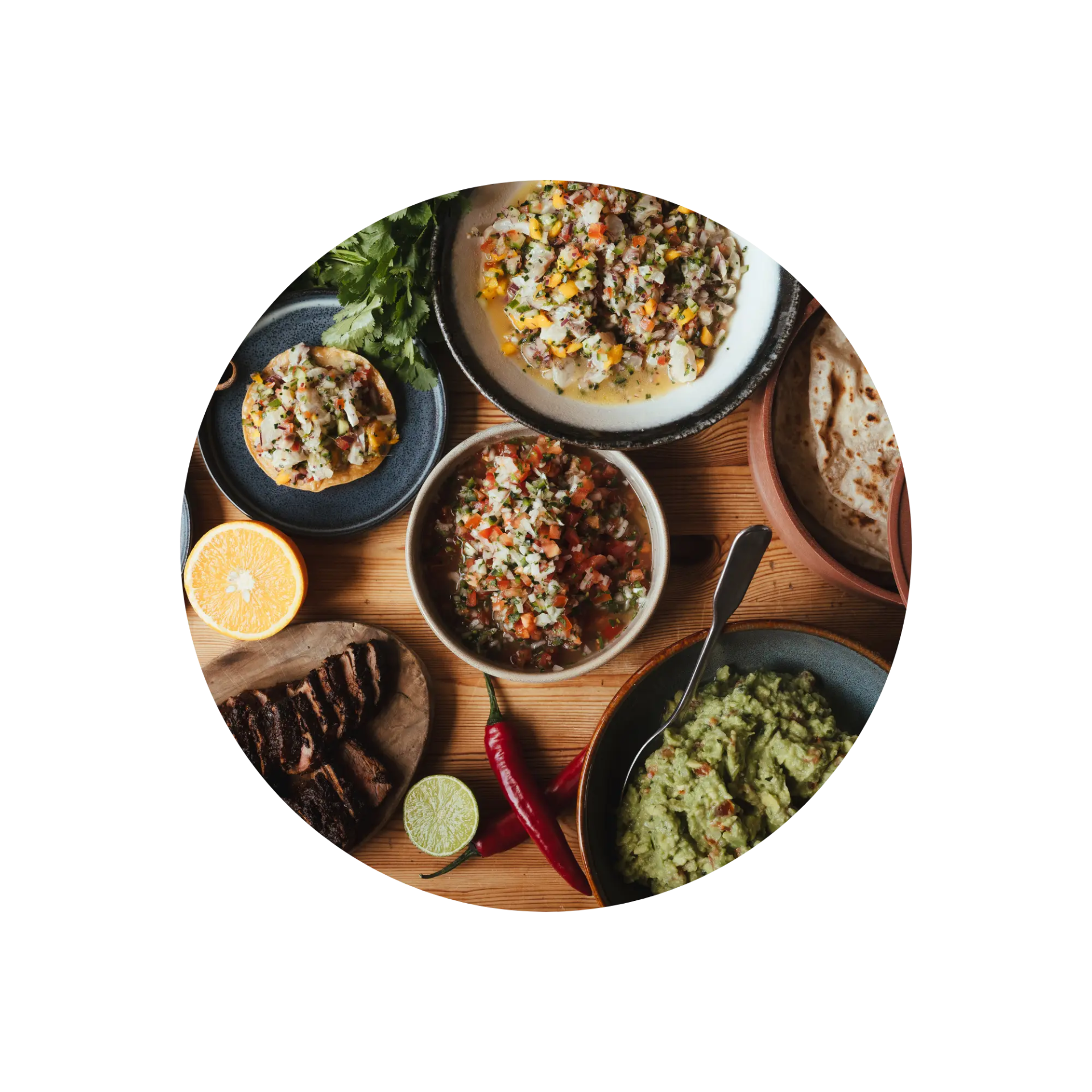 Mexican dishes with tortillas, guacamole, salsas, meat, and fresh citrus on a wooden table.