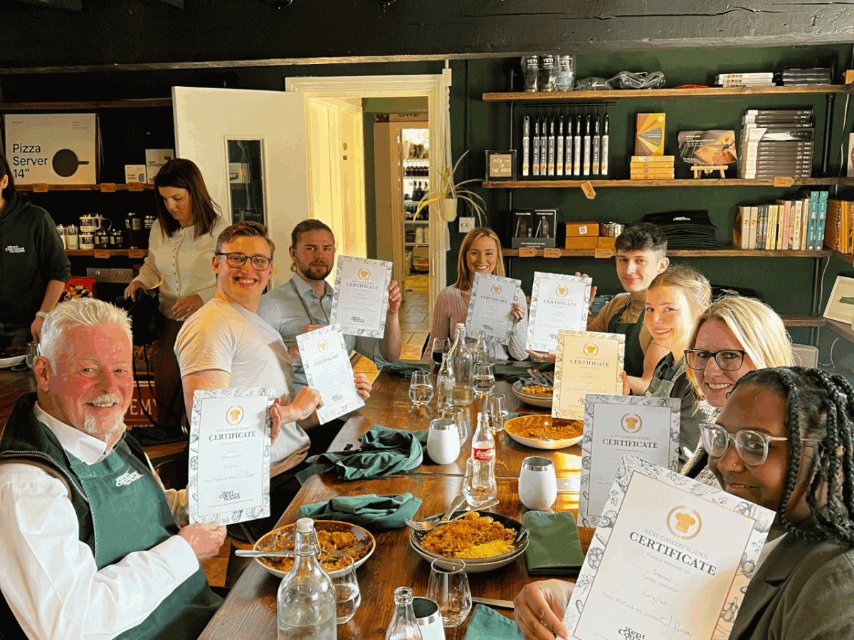 Group of people at a wooden table displaying certificates and food in a cozy room.