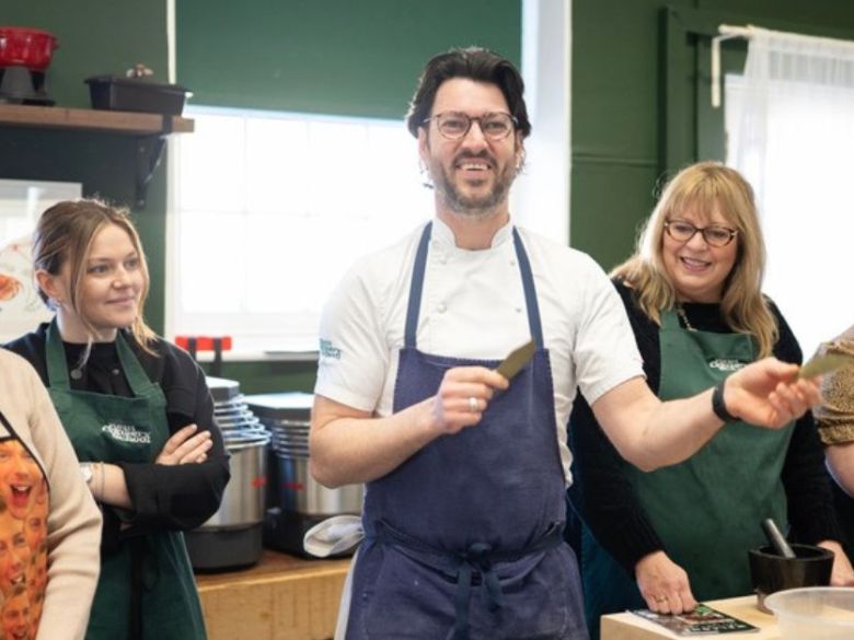 Group of people in a kitchen laughing, wearing aprons, with a man holding a utensil in the center.