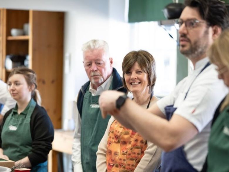 Group of people in aprons attending a cooking class, one person is gesturing while others listen.