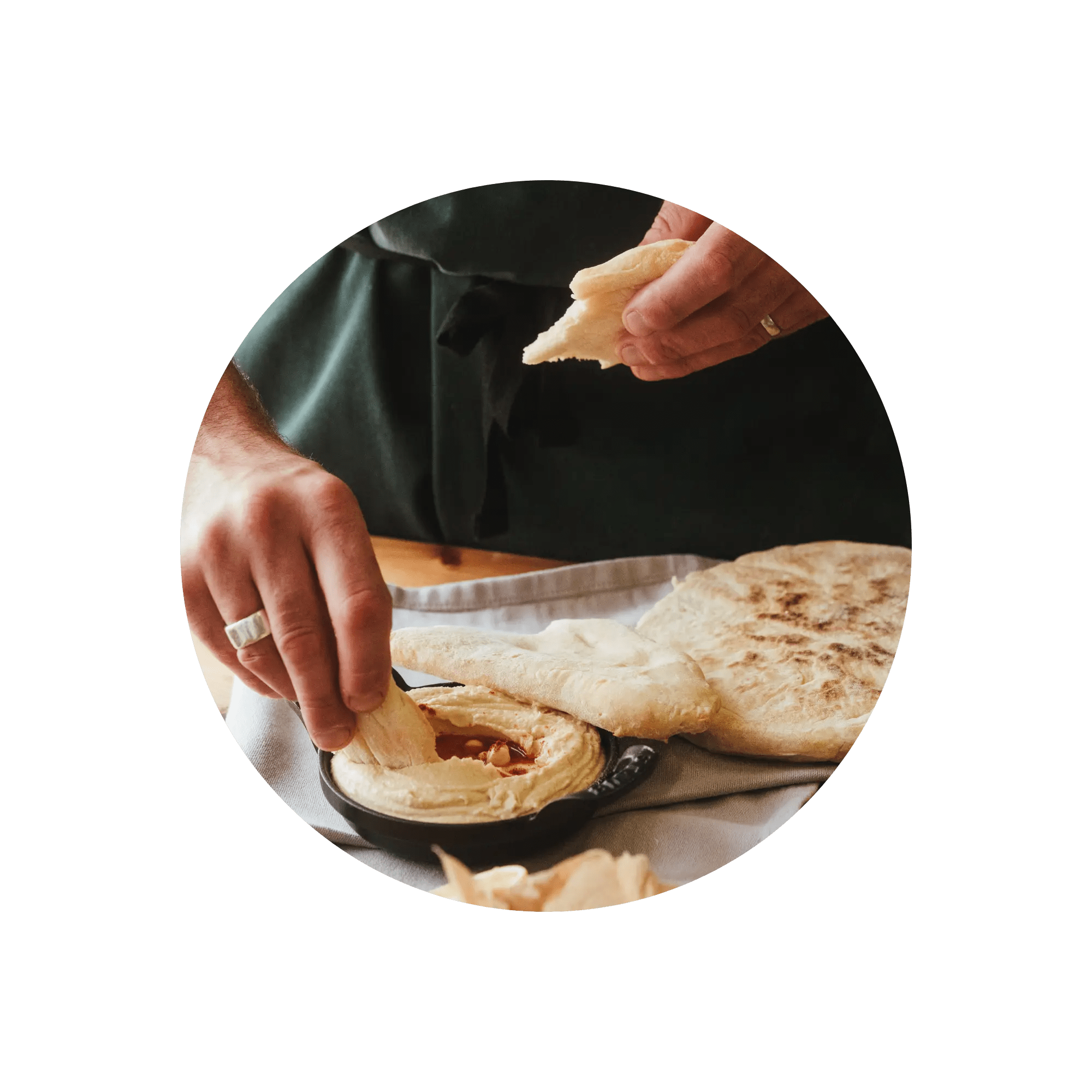 Hands dipping pita bread into a bowl of hummus on a table.