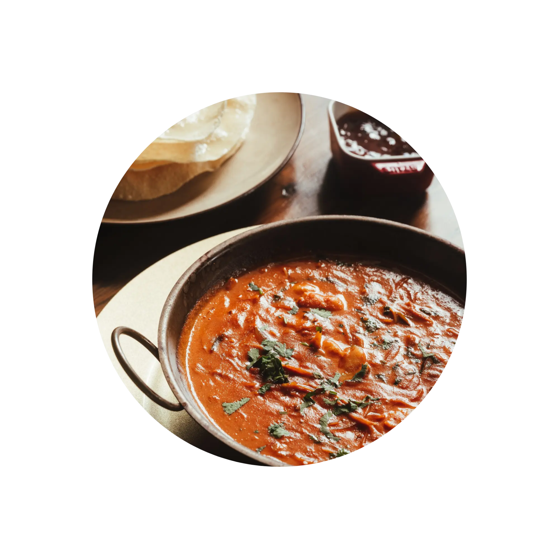 A bowl of curry with herbs, bread on side, on a wooden table.