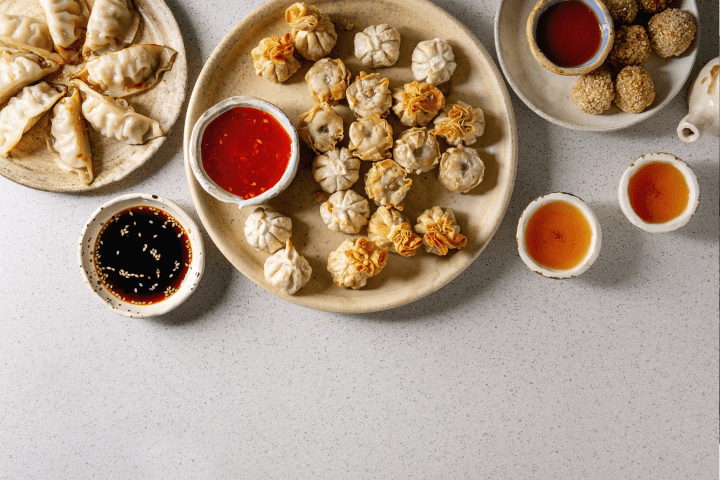 Assorted dumplings on plates with sauces and tea cups on a light surface.